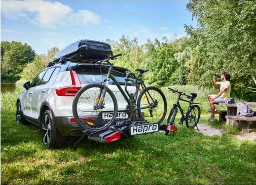 Hapro Atlas Premium Xfold II 3.0 montert p&aring; bil p&aring; gr&oslash;ntomr&aring;de, med sykler lastet og folk som slapper av ved et bord i bakgrunnen