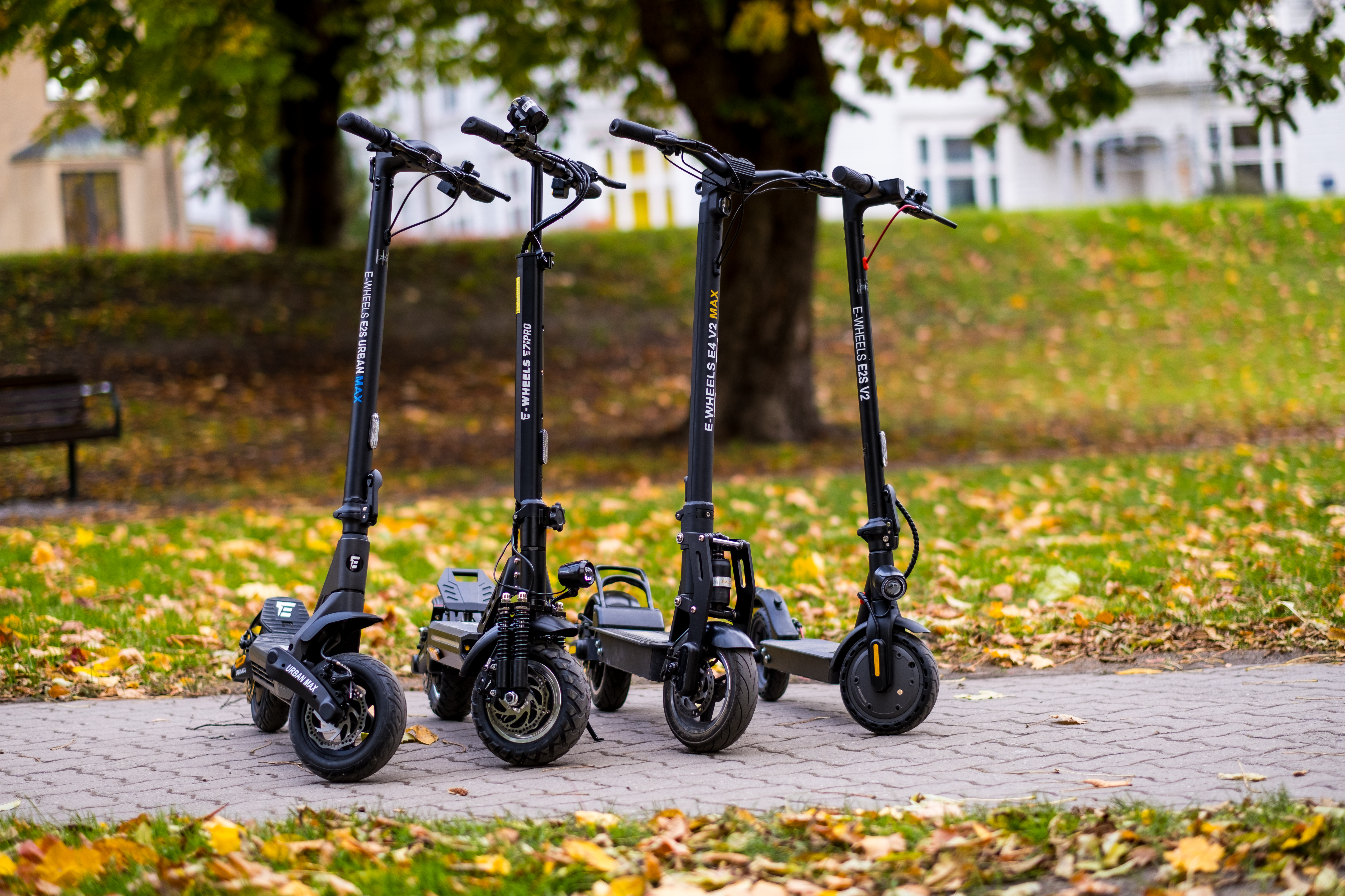 På bildet er det fire el-sparkesykler fra merker E-Wheels, b.la. E2S Urban Max, E7 Pro, E4 V2 og E2S V2. Bildet er tatt utendørs. Alle sparkesyklene er sorte. 